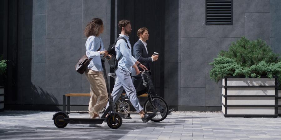 une femme en trottinette, un homme à côté de son vélo et un homme avec son café marchent sur un trottoir ensoleillé